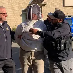 A woman in a full beekeeper suit is led away from a house by police officers.