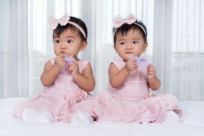 Twin babies wearing pink dresses and head bows, holding purple flowers, on a white bed
