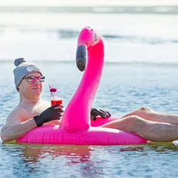 A man swimming in an ice hole in winter in Finland, floating on a pink inflatable flamingo with cocktail in hand. Vacation options, dreaming of summer.