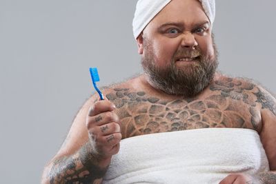 Close up of bearded tattooed man standing against the grey background and biting his lower lip while holding a toothbrush in right hand