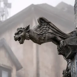 A photo of a medieval style grotesque in the form of a lion-headed winged create on the side of a building with a murky misty background showing a building.