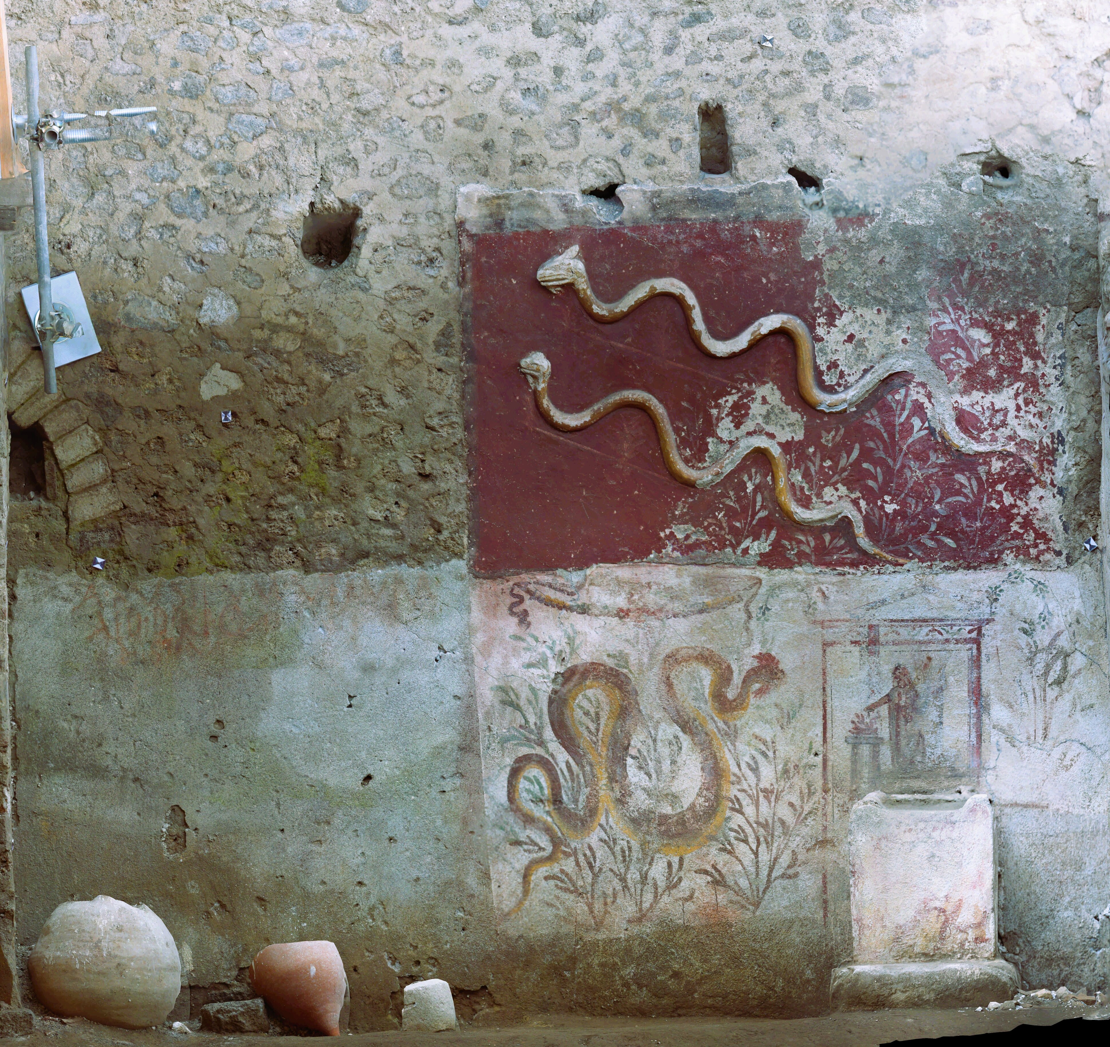 A wall of opus mixum is visible. A small portion of it is plastered on top in red with two plaster snakes and below a fresco of a snake and olives branches are visible. A small white altar is on the ground with behind the represantation of a person doing a ritual.