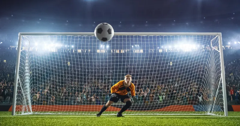 A football goalkeeper in front of the net as a football flies towards him 