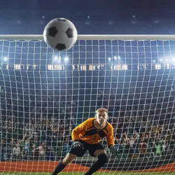 A football goalkeeper in front of the net as a football flies towards him 