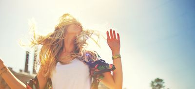 woman outside in bright sun with hands up and blonde hair flowing round her face