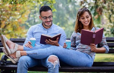 An overly happy couple sit and recline on a park bench while reading. The couple are intentionally or otherwise wearing the same colour denim jeans and shirts, while holding identical blue and white spotted coffee cups and are reading burgundy coloured books.