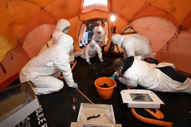 Six people in white forensic suits and masks are huddled around a hole in the earth as they excavate the human remains. The whole space is enclosed within an orang tent. There are other objects in the image, including an organ bucket that is nearly in the centre of the shot, as well as square shallow boxes that are being used to hold bone fragments. 