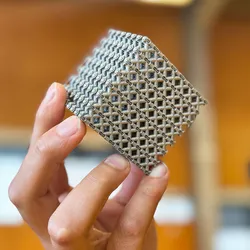 A photo of the new titanium cube being held in a man's hand. The photo is close up to the cube, showing its lattice arrangement and is the size of a Rubik's Cube. Faded in the background you can see the face of the man who holds it but his image is blurred. 