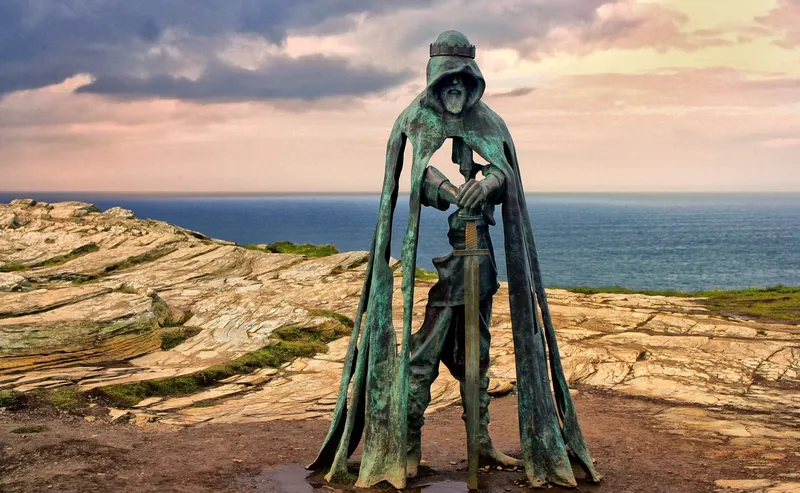 A photo of a bronze statue of King Arthur on a barren cliff side facing the sea. There are grey clouds in the background and a sunset sky. The sculpture shows an older man with a beard shrouded in a hood with a crown on the outside. The sculpture has been crafted so that parts of the body and cloak are missing, revealing the landscape behind it. The figure is wearing armour and is resting his gauntleted hands on the pummel of a sword that extends to the ground.  