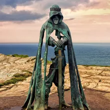 A photo of a bronze statue of King Arthur on a barren cliff side facing the sea. There are grey clouds in the background and a sunset sky. The sculpture shows an older man with a beard shrouded in a hood with a crown on the outside. The sculpture has been crafted so that parts of the body and cloak are missing, revealing the landscape behind it. The figure is wearing armour and is resting his gauntleted hands on the pummel of a sword that extends to the ground.  