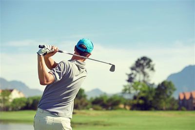 A photo of a man who has just swung a golf club standing on a golf course. The man's position shows the club over his left shoulder, suggesting he has just swung through after hitting the ball. He is wearing a light blue cap, grey t-shirt and white trousers. There is a pond blurred out to the left of the shot and there is a line of trees in the background.