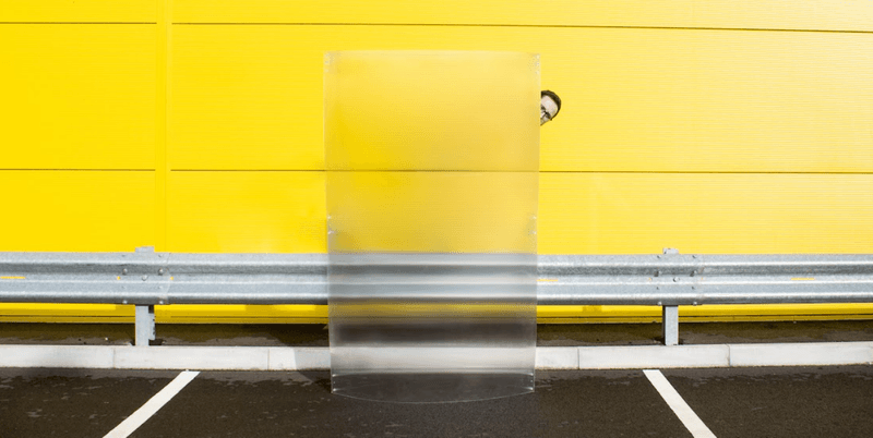 A man poking his out behind an invisibility shield in front of a yellow wall.