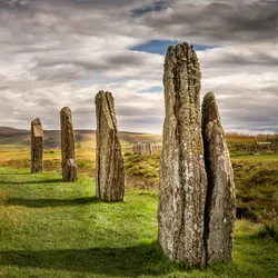 A close up photo of the standing stones at the Ring of Brodgar site in Orkney. The photo shows eight stones going into the distance as they mark out the circle of the structure. The stones sit on a band of grass which is surrounded by more unkept grasses. The sky is overcast with small slivers of blue.  