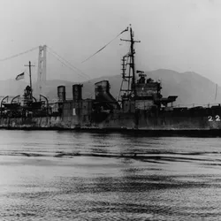 A black and white photo showing USS Stewart in the water with the Golden Gate bridge in the background. The water is calm and the vessel had two crew members standing on its bow. 