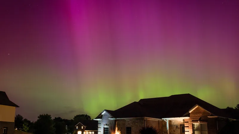 a few houses are seen at the bottom of the photo. In the night sky a stripe of green and a stripe of purple are visible, the aurora