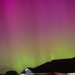 a few houses are seen at the bottom of the photo. In the night sky a stripe of green and a stripe of purple are visible, the aurora