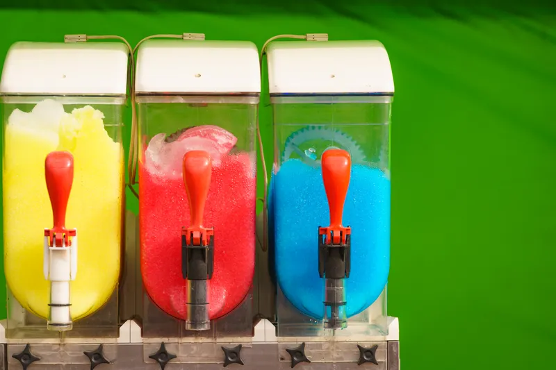 three machines containing slush drinks in yellow, red and blue, all lined up on a green background