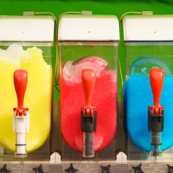 three machines containing slush drinks in yellow, red and blue, all lined up on a green background