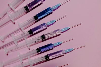 syringes arranged on a pink background; they're all facing towards the upper right corner and contain liquid that is either clear, pale blue, or pale purple