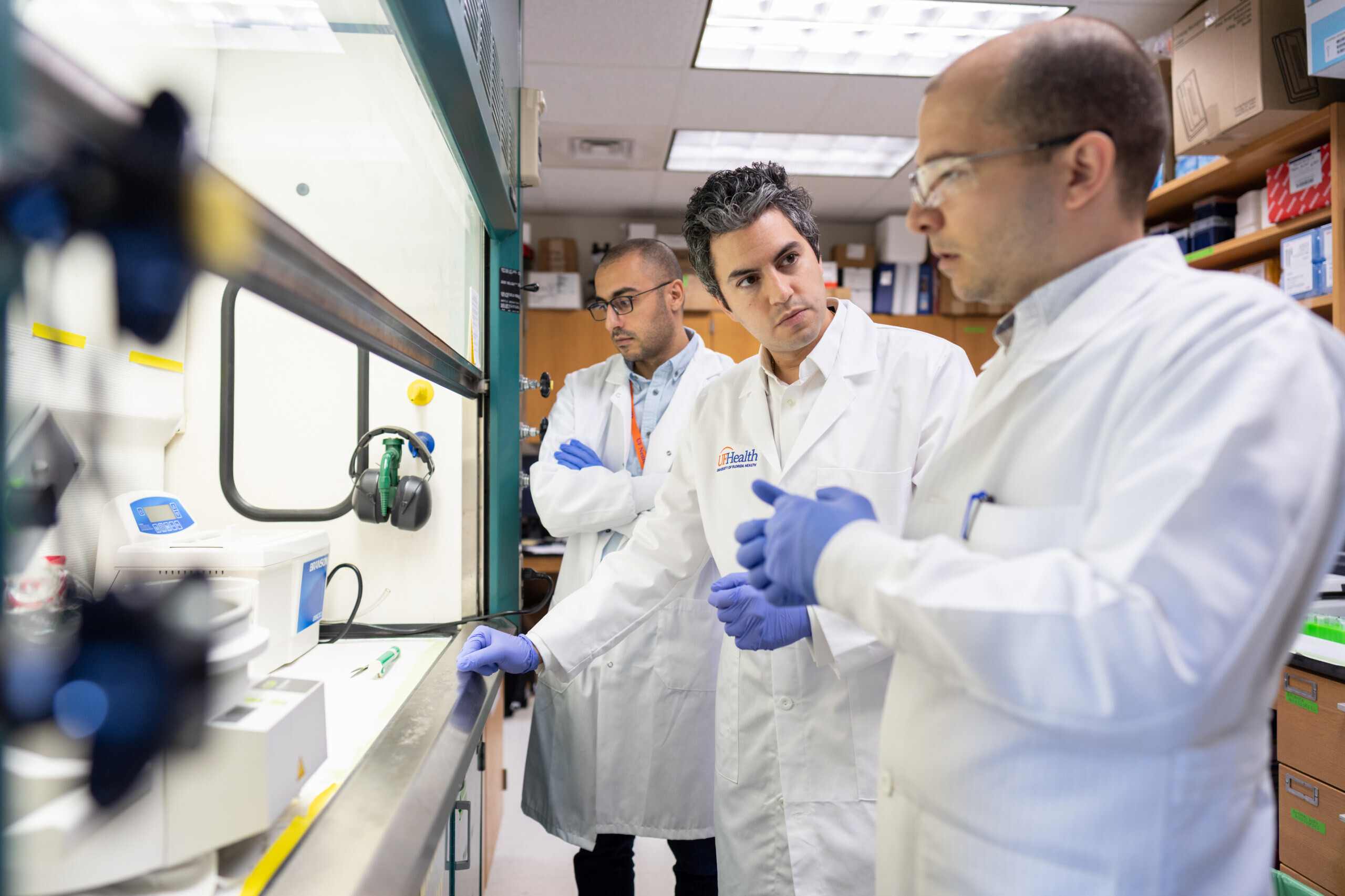 three male scientists wearing lab coats and gloves standing in front of a culture hood in a lab with lots of brown shelving units and cabinets behind them