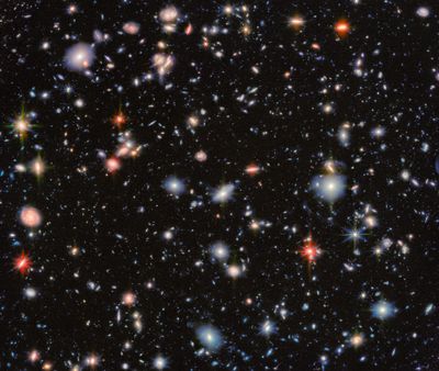An area of deep space with thousands of galaxies in various shapes and sizes on a black background. Most are circles or ovals, with a few spirals. More distant galaxies are smaller, down to being mere dots, while closer galaxies are larger and some appear to be glowing. Red and orange galaxies contain more dust or more stellar activity.