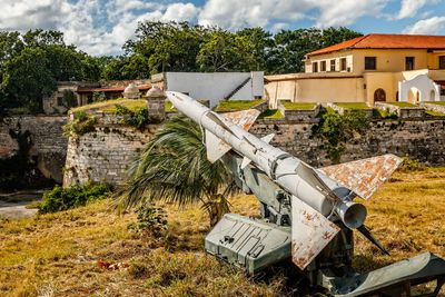 A photo of a rusted missile left out on what looks like a street edge in Cuba. The missile is facing away from the viewer, it's tail fins heavily rusted. There is a small palm tree right in front of it and a few meters away a brick wall, which may be the edge of a battlement. In the background a large building is visible with tanned stone and red clay roof tiles. 