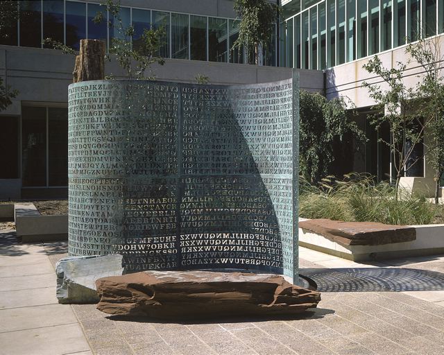 A photo showing the Kryptos sculpture on site at the CIA headquarters. It looks like a large metal scrolling being unfolded from an upright piece of petrified wood (on the left). In front of it is a brown rock bench. The sunlight shines through portions of the letters carved into its surface, which projects them on the floor in its shadow. Behind the sculpture, a seating area is visible with some grasses and slim trees and there is a large building in the background.
