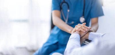 A cropped photo showing a doctor or nurse in green scrubs holding the hand of a patient sitting in a chair. The photo is focused on then hands and shows the torso and upper legs of the doctor from the perspective positioned just over the patient's left shoulder. 
