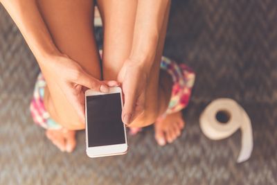 aerial view of woman using smartphone while on the toilet; only her legs are visible, and there is a roll of toilet paper on the floor on the right of the image