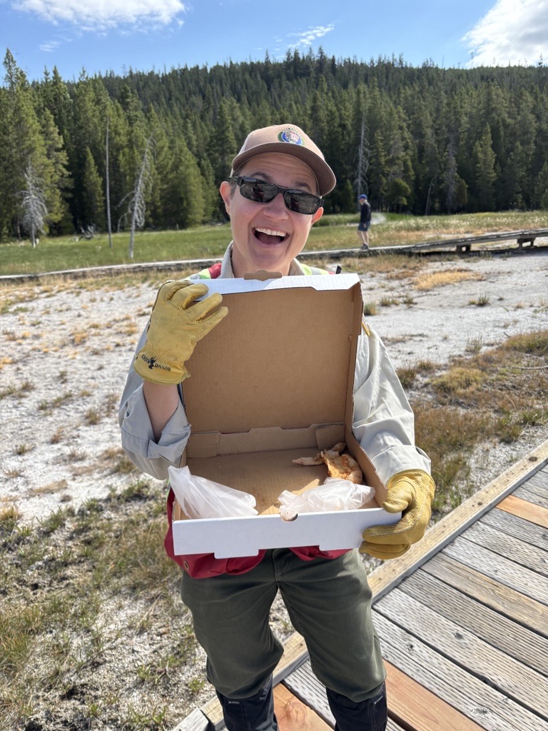 Yellowstone National Park Geology Program volunteer Tara Cross smiles and shows off a pizza box found on Geyser Hill in Upper Geyser Basin