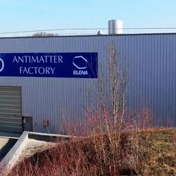a photo of the antimatter factory from the outside. It is a generic grey warehouse with a big blue sign that says antimatter factory and a large grey shutter.