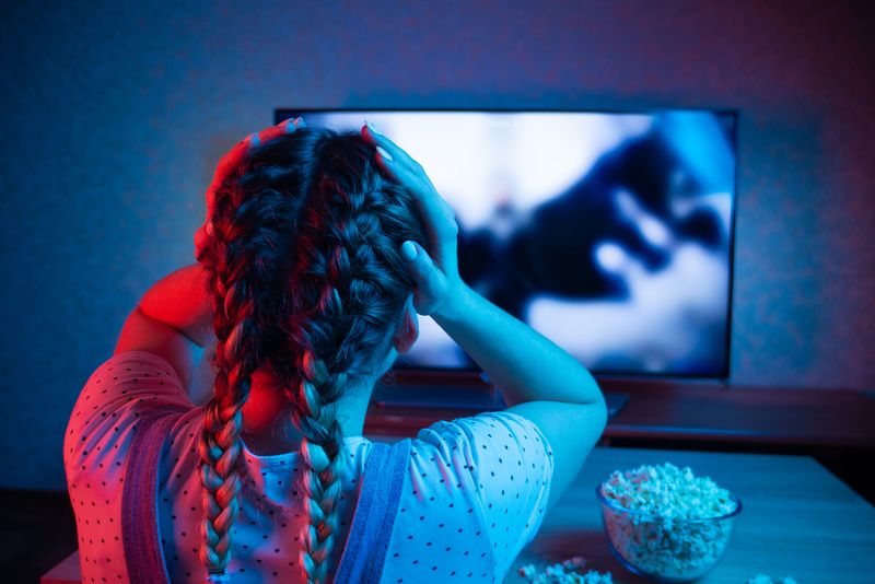 young woman pictured from behind holding her hands to her head while she watches a movie on TV, the lighting is dark with red and purple. There's a bowl of popcorn on the table in front of her. She has two braids in her hair and is wearing dungarees and a polka dot t-shirt.