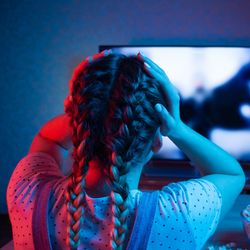 young woman pictured from behind holding her hands to her head while she watches a movie on TV, the lighting is dark with red and purple. There's a bowl of popcorn on the table in front of her. She has two braids in her hair and is wearing dungarees and a polka dot t-shirt.