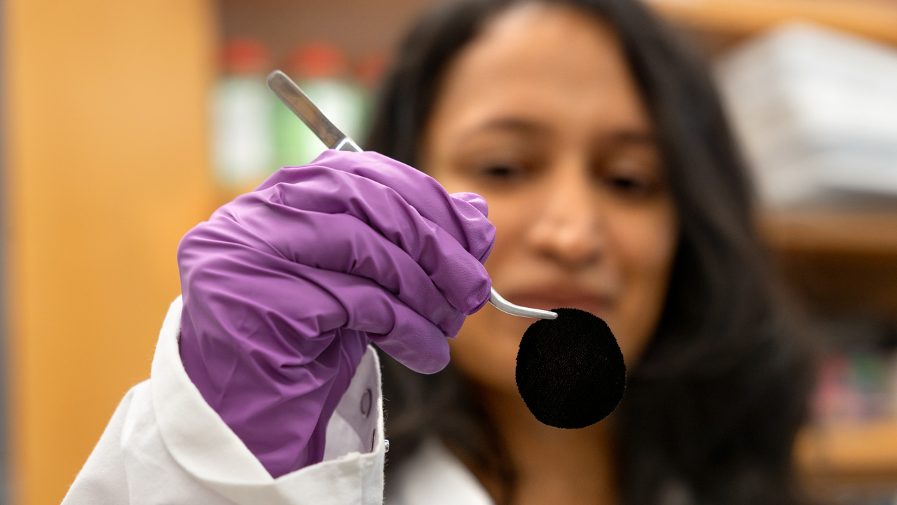out of focus researcher holding a circle of very dark fabric in a pair of tweezers, while wearing a lab coat and purple gloves