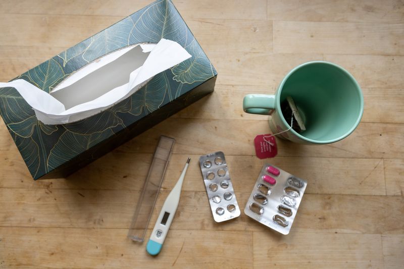 aerial shot of table with box of tissues, digital thermometer, partially empty blister packs of pills, and a mug with a teabag