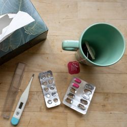 aerial shot of table with box of tissues, digital thermometer, partially empty blister packs of pills, and a mug with a teabag