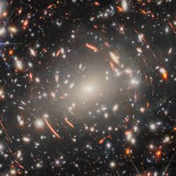 A field of galaxies in space, dominated by an enormous, bright-white elliptical galaxy that is the core of a massive galaxy cluster. Many other elliptical galaxies can be seen around it. Also around it are short, curved, glowing red lines, which are images of distant background galaxies magnified and warped by gravitational lensing. A couple of foreground stars appear large and bright with long spikes around them.