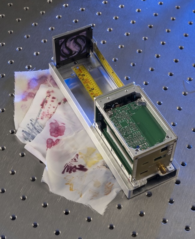 Examples of the fabric painted with bacterial dyes sit beside the Spinning Around SpinnyONE satellite which will carry it into space next year