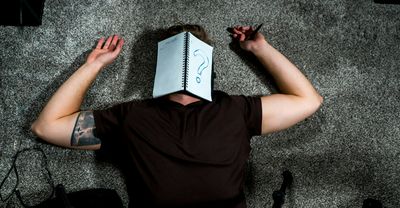 man sleeping on the floor with his face covered by an open notebook, with a large question mark written on one of the pages