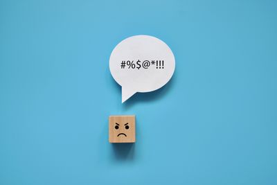 wooden block with angry face on blue background with a speech bubble containing symbols to represent profanity