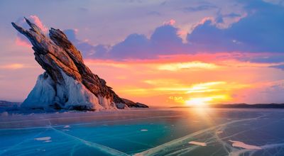 the lake is frozen with a big rock formation sticking out and the sun rising in the distance