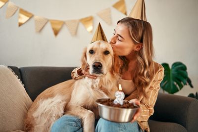 Young happy woman holding birthday cake, celebrating her pet dog's birthday, hugging and having fun with her pet while sitting in the living room at home.
