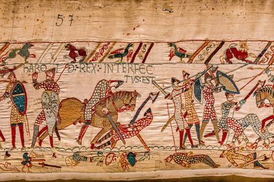 A section of the Bayeux Tapestry depicting the scene where Harold II was shot in the eye by an arrow. Harold appears on the left as a two dimensional figure reaching up to grab the arrow shaft in his head. Immediately next to him is horseman cutting down a warrior with an axe, and beyond them is a group of soldiers with spears.