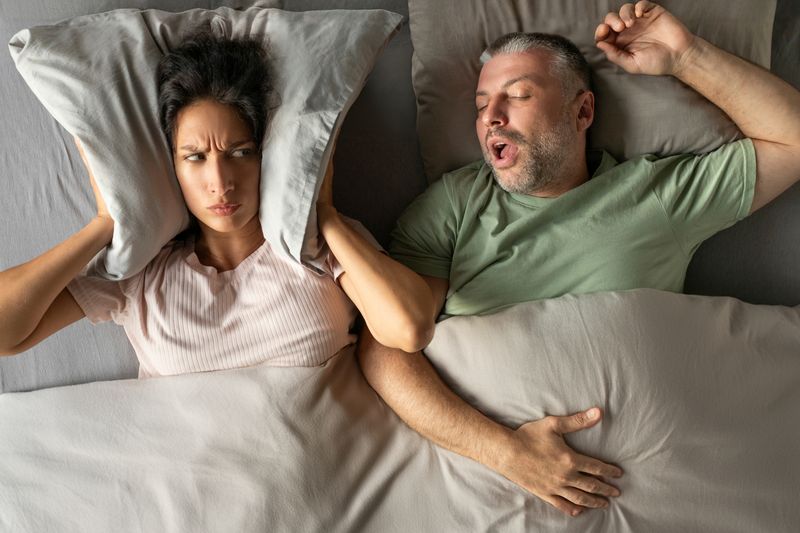 Unhappy tired woman covering her ears with pillow, can not handle loud snore of her husband, top view shot
