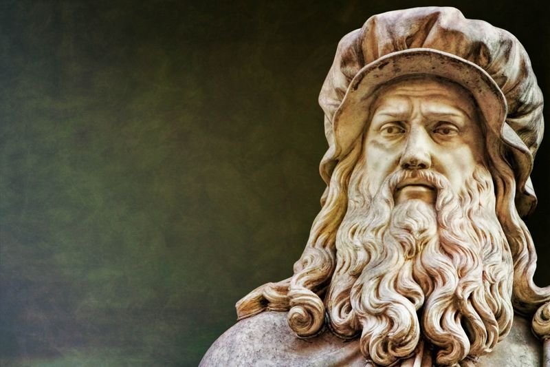 A close up of a statue of Leonardo da Vinci. The photo shows the statue's head sporting a large beard that comes down to the artist's chest, as well as his shoulder length hair partially covered by his iconic cap. 
