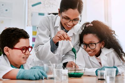 A photo of a science teacher making a demonstration with some green goo substance in a bowl. She is wearing a white coat and holding a stick with the substance running off it into a bowl on the table below here. She has to children in white coats and rubber gloves leaning in beside her to get a better look at the substance.