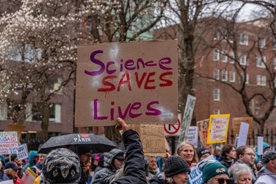 A photo showing a protest where someone is holding up a large square of cardboard with the words "Science Saves Lives" written on it is purple and red color. The rest of the photo shows the heads of protests, many holding up plaques and many wearing raincoats and hats to protect them from the winter weather. 
