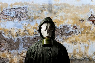 A photo of someone wearing a gas mask and a green plastic-like suit covering their body. They are standing looking directly at the camera with their back to a weathered, stained wall.