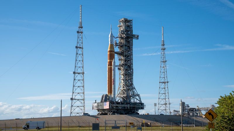 The rocket, orange, with two white boosters and a white top capsule, stands at the launchpad.