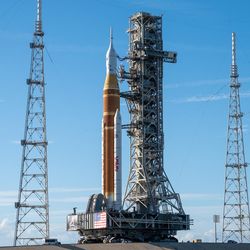 The rocket, orange, with two white boosters and a white top capsule, stands at the launchpad.
